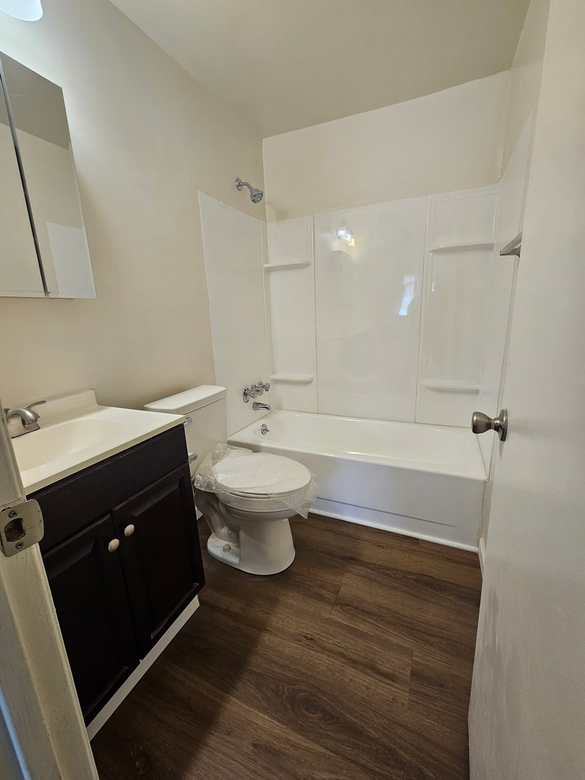 A bathroom at Village Green Apartments with a white tub and shower surround, a dark vanity with a mirrored cabinet, and plank flooring