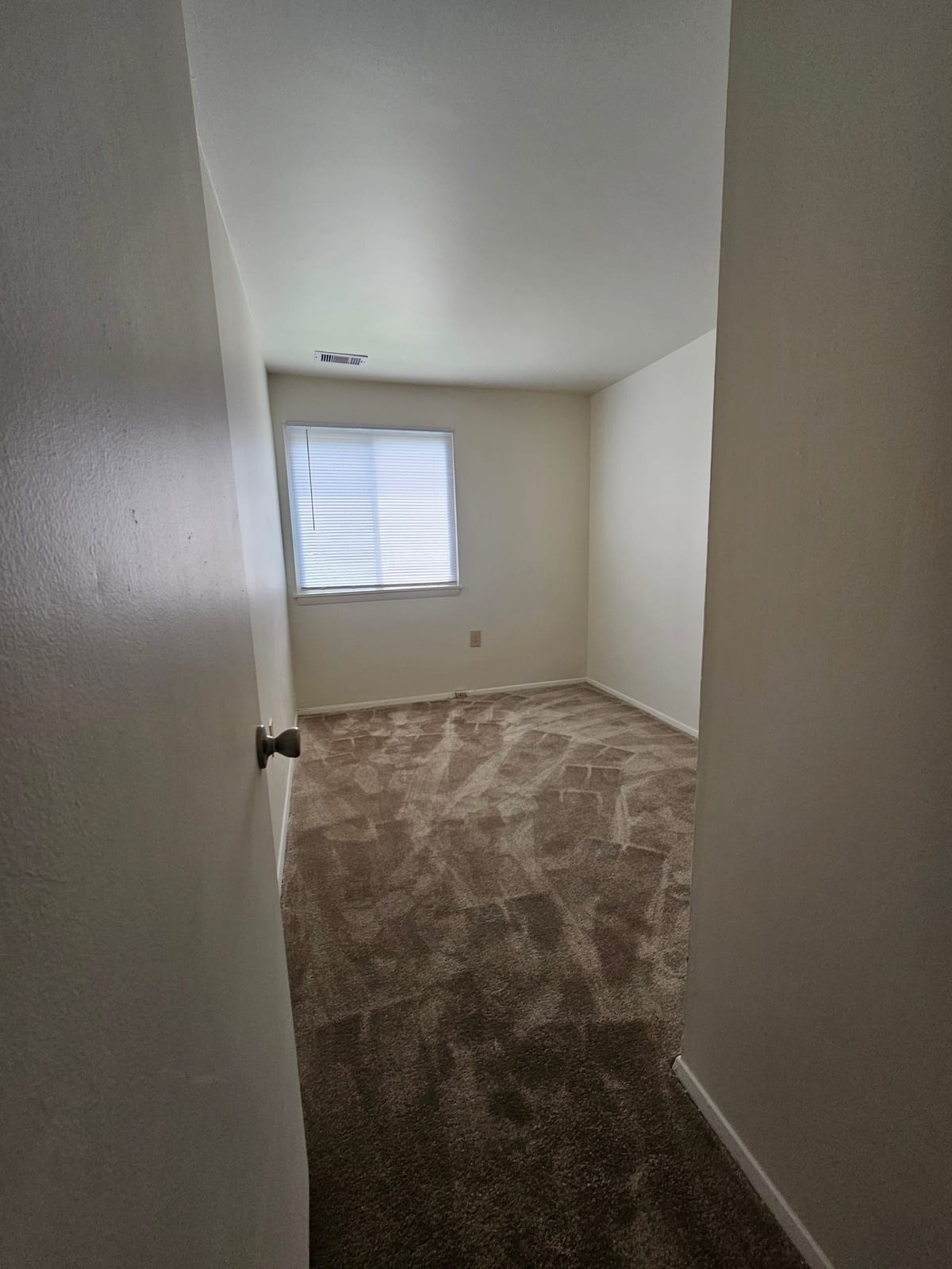 A second carpeted bedroom at Village Green Apartments with a blind-covered window, seen through the open doorway