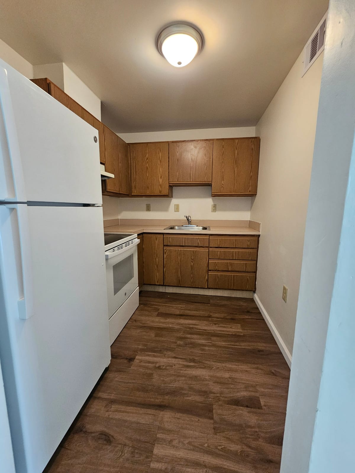 A kitchen at Village Green Apartments with oak cabinets, a white electric range and refrigerator, a stainless sink, and wood-look plank flooring
