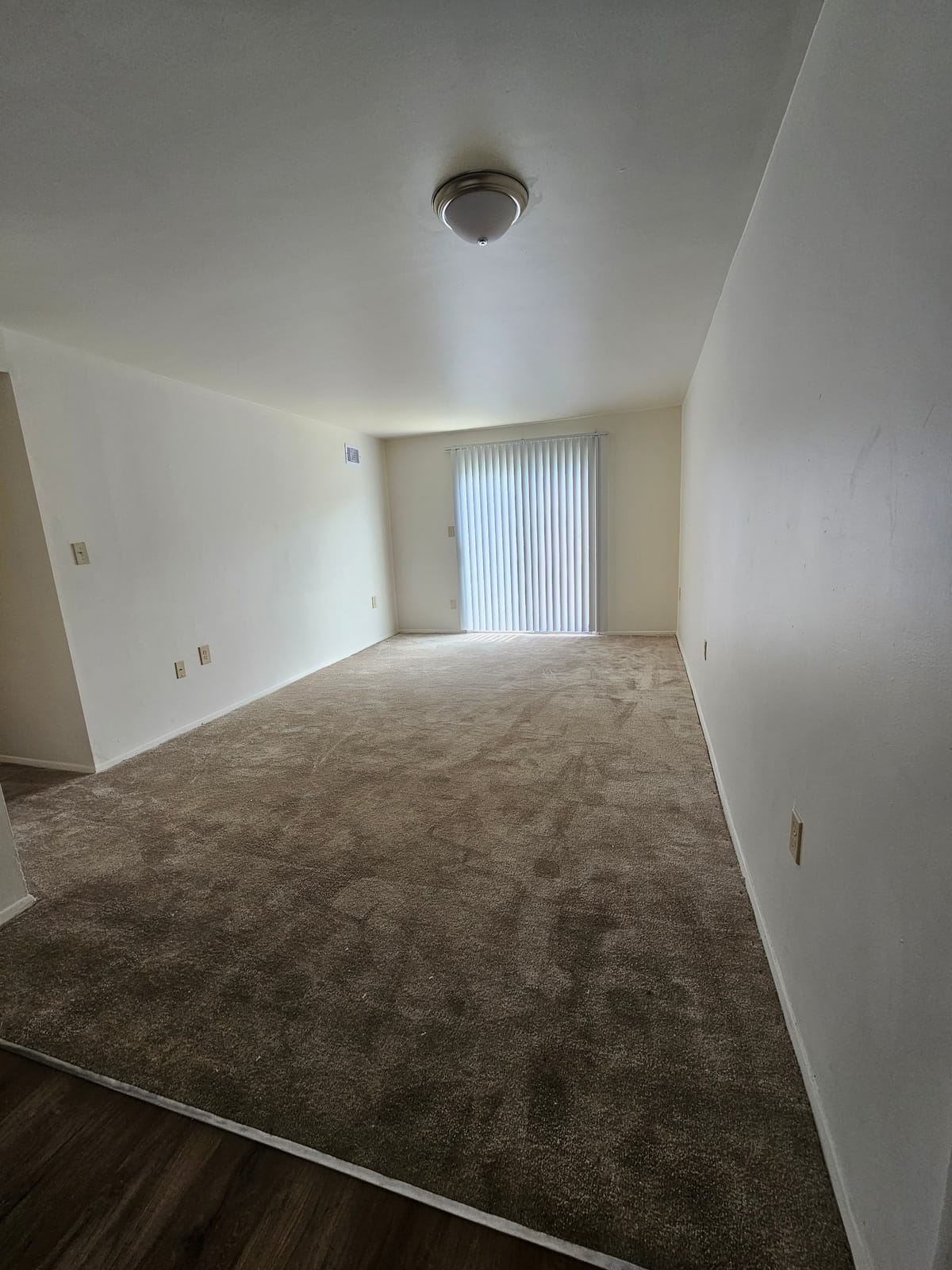 A carpeted living room at Village Green Apartments with a sliding glass door covered by closed vertical blinds