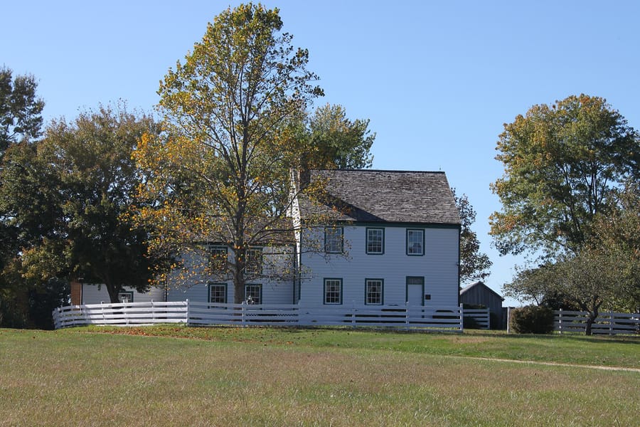 The white clapboard farmhouse of the Dr. Samuel A. Mudd House Museum behind a white fence, framed by autumn trees