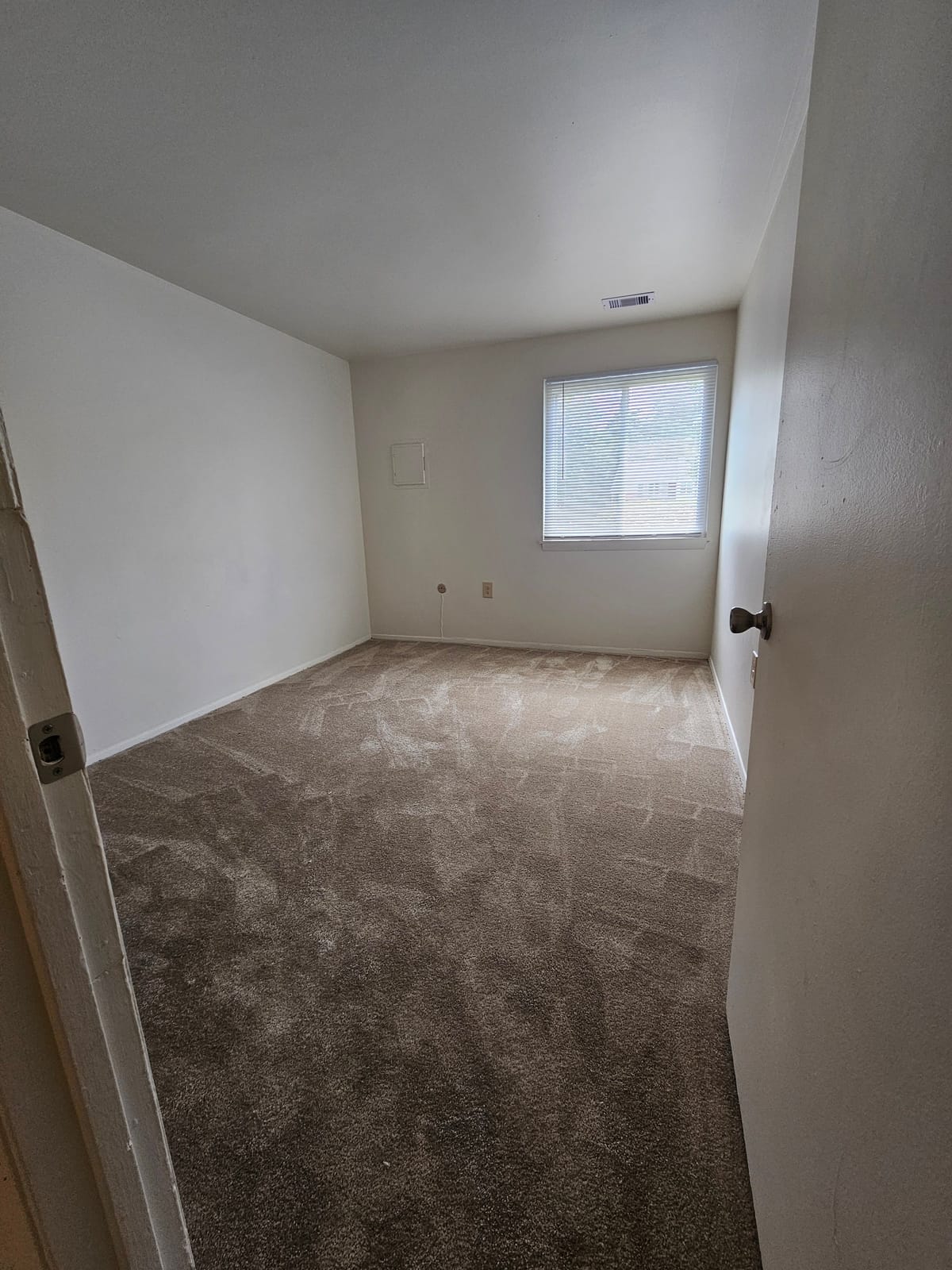 A carpeted bedroom at Village Green Apartments with a blind-covered window, seen from the doorway